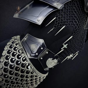May include: Close-up of black armor with silver accents. The armor features a shoulder plate, chainmail, and a decorative belt with silver coin-like embellishments. Silver spikes and cross-shaped charms hang from the chainmail, adding detail to the medieval-inspired design.