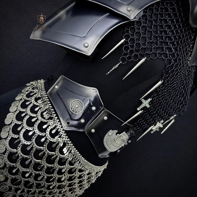 May include: Close-up of black armor with silver accents. The armor features a shoulder plate, chainmail, and a decorative belt with silver coin-like embellishments. Silver spikes and cross-shaped charms hang from the chainmail, adding detail to the medieval-inspired design.
