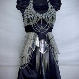 K&ouml;nnte beinhalten: Ein silbernes Kettenhemd-Oberteil und ein Metallpanzer-Rock-Set, das auf einer schwarzen Schaufensterpuppe ausgestellt ist. Die R&uuml;stung umfasst ein Kettenhemd-Haltertop, einen Metallg&uuml;rtel und eine geschichtete Metall-H&uuml;ftr&uuml;stung mit herabh&auml;ngenden Ketten. Das Logo "STEEL HERITAGE BATTLE ARMORS" ist sichtbar.