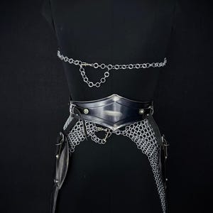 May include: A black mannequin displays a silver chainmail and leather battle armor set. The armor includes a chainmail skirt, a leather belt with metal accents, and a chainmail top. The Steel Heritage logo is visible in the upper left corner.