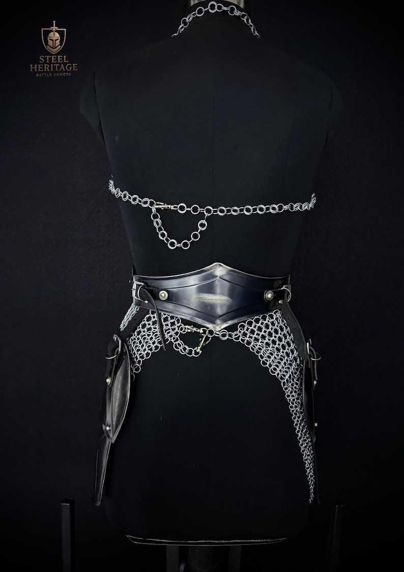 May include: A black mannequin displays a silver chainmail and leather battle armor set. The armor includes a chainmail skirt, a leather belt with metal accents, and a chainmail top. The Steel Heritage logo is visible in the upper left corner.
