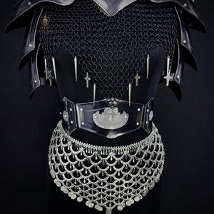 May include: Black medieval armor set featuring a chainmail shirt, shoulder pauldrons, and a studded belt with a decorative buckle. The armor is adorned with silver crosses and pendants, and a silver chainmail skirt.