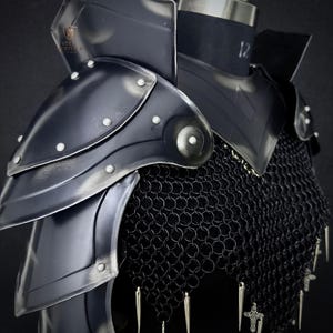 May include: Close-up of a black steel shoulder armor set with riveted accents and a chainmail underlayer. The armor is displayed on a mannequin, showcasing its design and construction. The Steel Heritage logo is visible.