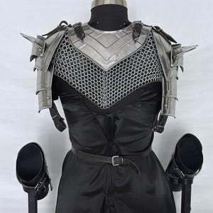 May include: A medieval-style steel armor set, including a chest plate, shoulder guards, and chainmail, displayed on a mannequin. The armor is silver-colored and features pointed shoulder spikes. The mannequin is dressed in a black outfit with a leather belt.
