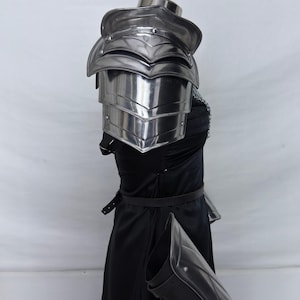 May include: Polished steel shoulder armor with layered plates, part of a medieval battle armor set. The armor is displayed on a black dress, with a matching arm guard. The Steel Heritage logo is visible in the upper left corner.