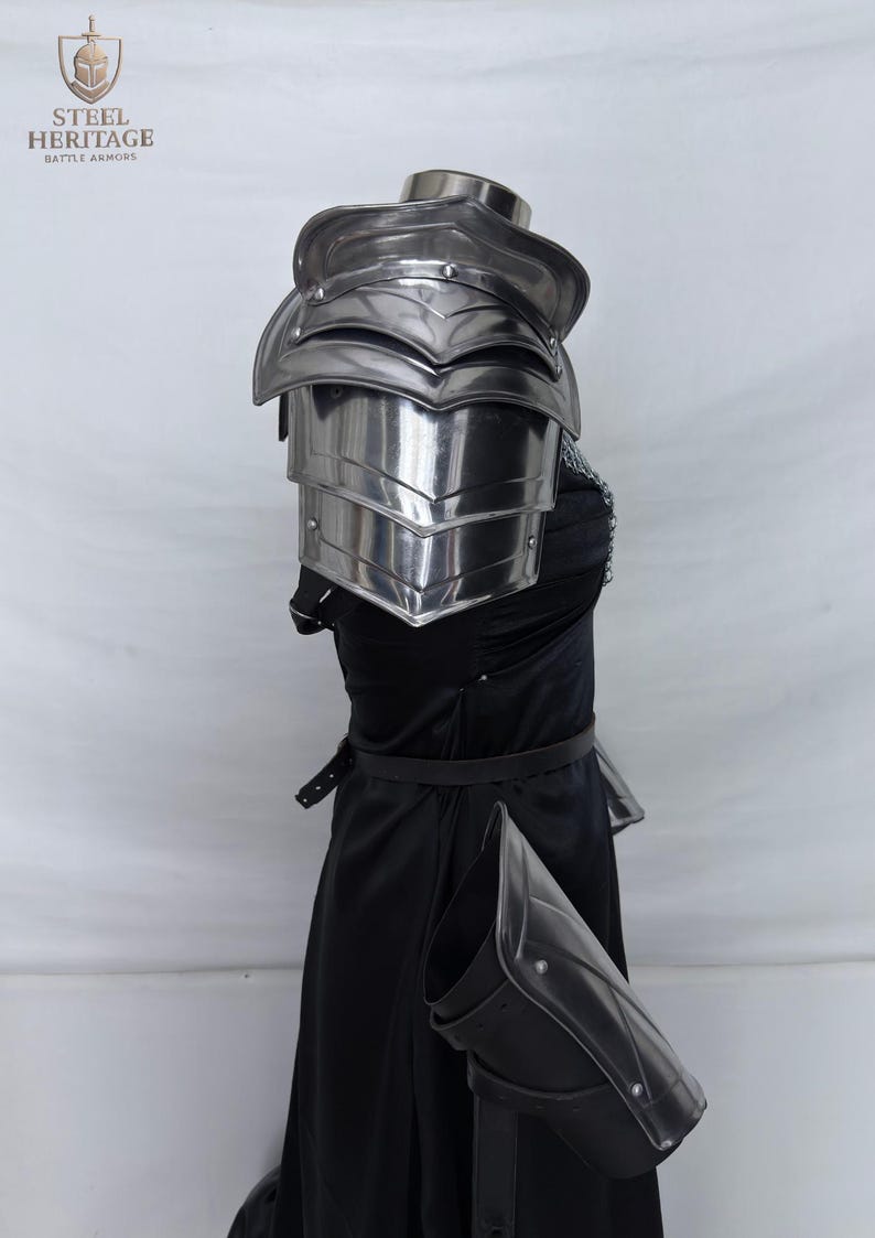 May include: Polished steel shoulder armor with layered plates, part of a medieval battle armor set. The armor is displayed on a black dress, with a matching arm guard. The Steel Heritage logo is visible in the upper left corner.