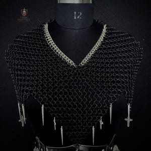 May include: Black chainmail armor with a V-neck design, accented with silver trim and dangling silver spikes and crosses. The armor is displayed on a black mannequin. The Steel Heritage logo is visible in the upper left corner.
