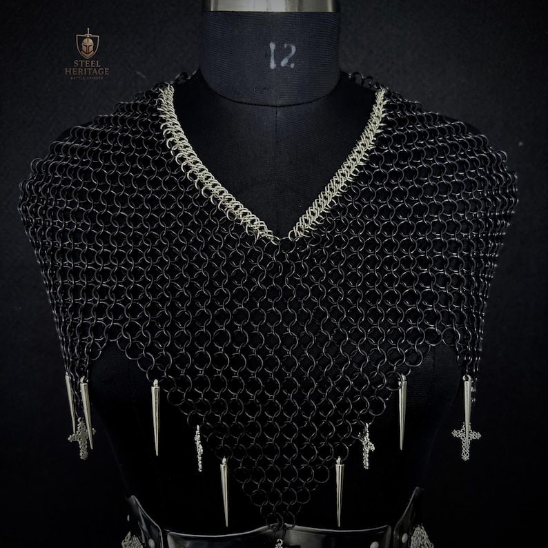 May include: Black chainmail armor with a V-neck design, accented with silver trim and dangling silver spikes and crosses. The armor is displayed on a black mannequin. The Steel Heritage logo is visible in the upper left corner.