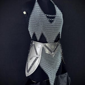 May include: A silver chainmail top and belt set displayed on a black mannequin. The top is a halter style with a chainmail bodice. The belt is silver with decorative accents and a chainmail skirt.