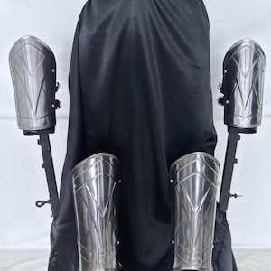 May include: A set of silver-colored arm and forearm armor, displayed against a black backdrop. The armor pieces have a geometric design and are mounted on black stands. The overall aesthetic suggests a medieval or fantasy theme.