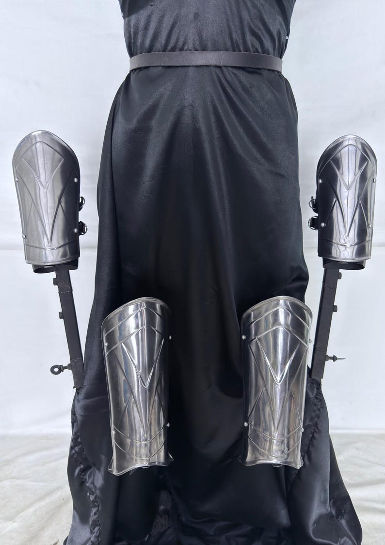 May include: A set of silver-colored arm and forearm armor, displayed against a black backdrop. The armor pieces have a geometric design and are mounted on black stands. The overall aesthetic suggests a medieval or fantasy theme.