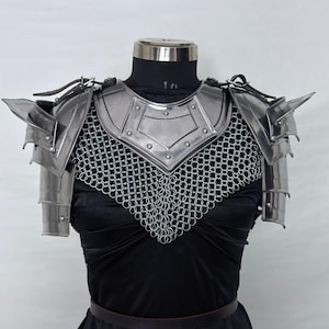 May include: Polished steel battle armor, including shoulder pauldrons and a chainmail collar, displayed on a mannequin. The armor is designed with sharp angles and riveted plates. The Steel Heritage logo is visible in the upper left corner.