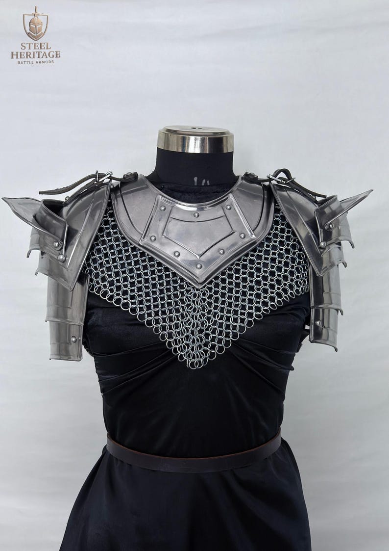May include: Polished steel battle armor, including shoulder pauldrons and a chainmail collar, displayed on a mannequin. The armor is designed with sharp angles and riveted plates. The Steel Heritage logo is visible in the upper left corner.