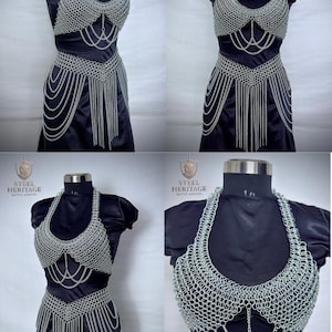 K&ouml;nnte beinhalten: Ein silbernes Kettenhemd- und G&uuml;rtel-Set, das auf einer schwarzen Schaufensterpuppe pr&auml;sentiert wird. Das Oberteil hat ein Neckholder-Design und aufwendige Kettendetails. Der G&uuml;rtel hat einen kaskadierenden Kettenfransen. Das Steel Heritage-Logo ist sichtbar.
