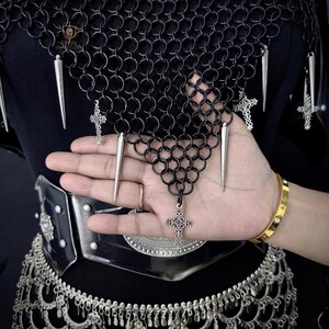May include: Black chainmail necklace with silver spikes and cross pendants. The necklace is made of small, interlocking metal rings. The design includes a black belt and silver trim. The overall aesthetic is gothic and edgy.