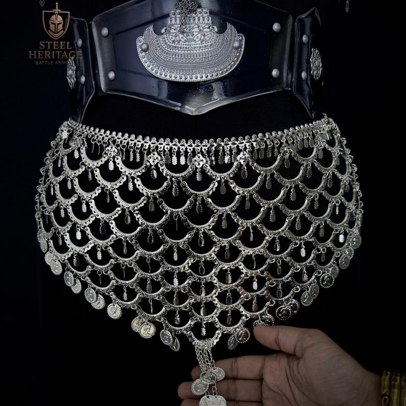 May include: A silver-colored battle armor piece with a decorative design. The armor features a wide belt with ornate detailing and a lower section with a scale-like pattern and dangling coins. The Steel Heritage logo is visible.