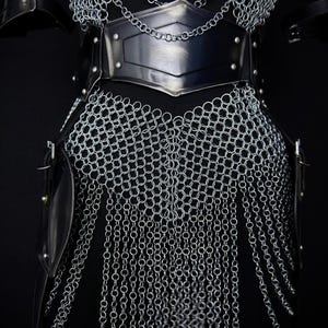 May include: Close-up of a medieval chainmail armor set. The armor features a silver chainmail design with a black leather belt and accents. The Steel Heritage logo is visible in the upper left corner. The armor is designed to protect the torso.
