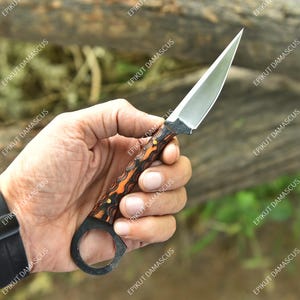 May include: A small, sharp knife with a silver blade and an orange and black patterned handle. The knife has a ring at the base of the handle. The text "EPIKUT DAMASCUS" is repeated in the background.