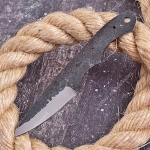 May include: A fixed blade knife blank featuring a dark, textured, forged finish on the upper portion of the blade and handle, with a polished, silver-colored sharpened edge. The handle section includes three circular holes for mounting scales.