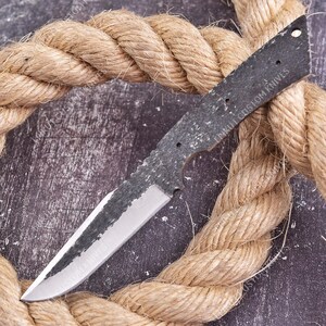 May include: A fixed blade knife blank with a dark, textured, forged finish on the spine and handle area, transitioning to a polished, sharp steel edge. The handle section features three pre-drilled holes and the text PAKUL CUSTOM KNIVES.