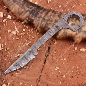 May include: A Damascus steel knife with a unique design. The knife features a pointed blade and a handle with a circular opening. The blade and handle have a patterned, textured appearance. The knife is displayed on a wooden surface.