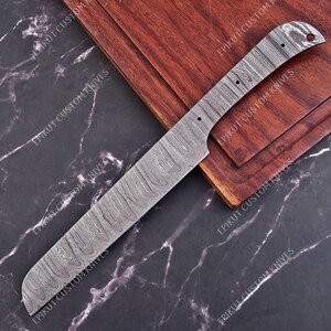 May include: A Damascus steel knife blade with a wavy pattern, resting on a wooden cutting board. The blade has a rectangular shape with a curved handle end. The background is a dark marble surface. The text "EPIKUT CUSTOM KNIVES" is visible.