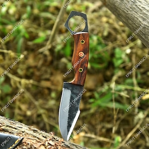 May include: A handcrafted knife with a dark, textured blade and a wooden handle. The handle features brass rivets and a decorative emblem. The knife has a unique hexagonal hole at the top of the handle. The blade is approximately 4 inches long.