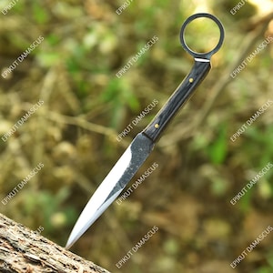 May include: A handmade knife featuring a dark wooden handle and a circular finger hole. The silver blade has a sharp point. The knife is displayed against a blurred natural backdrop.