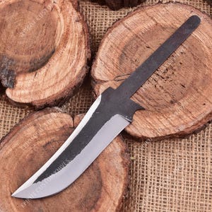 May include: A metal knife blade blank with a dark, textured finish on the spine and a polished, sharpened edge. The blade features a pointed tip and a long, narrow tang for handle attachment. The text EPIKUT CUSTOM KNIVES is visible on the blade.