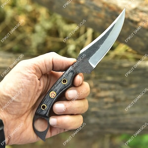 May include: A handmade knife with a curved, silver blade and a dark wooden handle. The handle features brass accents and a finger ring. The blade is held in a hand against a blurred natural background. The text "EPIKUT DAMASCUS" is visible.