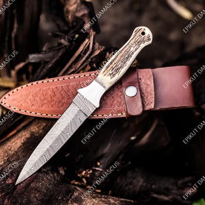 May include: A Damascus steel dagger with a patterned blade and a bone handle rests on a brown leather sheath. The sheath is embossed with a decorative pattern. The dagger is a hunting or utility knife.