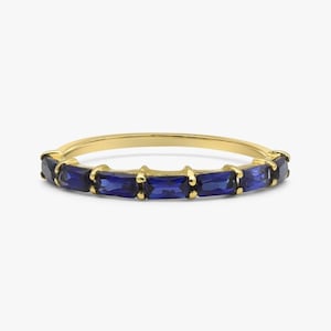 Könnte beinhalten: Ein goldener Bandring mit einer Reihe rechteckiger, tiefblauer Edelsteine. Die Edelsteine sind gleichmäßig um den Ring angeordnet und bilden ein durchgehendes Muster. Der Ring hat eine polierte, reflektierende Oberfläche.