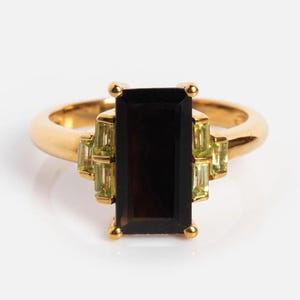 Könnte beinhalten: Ein goldener Ring mit einem großen, rechteckigen, dunkelbraunen Edelstein im Mittelpunkt. Der Ring ist mit kleineren, hellgrünen Edelsteinen auf beiden Seiten des Hauptsteins verziert. Das Band ist schlichtes, poliertes Gold.
