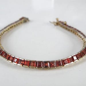 Pode incluir: Uma pulseira dourada com uma série de pedras preciosas vermelhas escuras de corte quadrado. As pedras são colocadas juntas, criando uma linha contínua de cor. A pulseira é projetada com um estilo clássico e elegante.