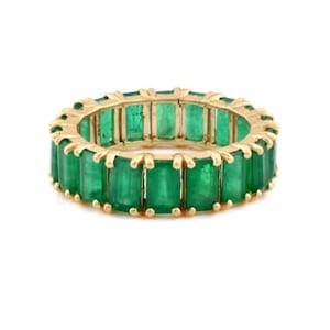 Könnte beinhalten: Ein goldener Ring mit einem durchgehenden Band aus smaragdgrünen Edelsteinen im Smaragdschliff. Die Fassung des Rings ist in einem warmen Goldton gehalten, und die Smaragde sind eng aneinandergereiht, was ein volles, lebendiges Aussehen erzeugt.