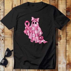 May include: Black t-shirt featuring a pink ghost design with a bow, holding a pink ribbon and a jack-o'-lantern. The ghost is covered in pink ribbons and a checkered pattern, symbolizing breast cancer awareness.
