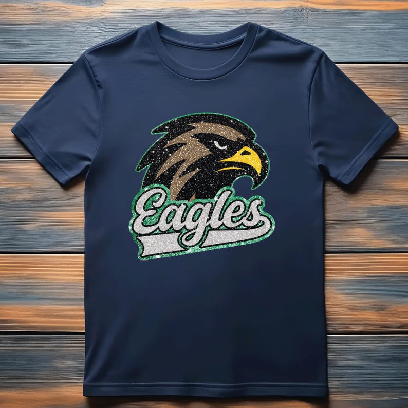 K&ouml;nnte beinhalten: Marineblaues T-Shirt mit einem glitzernden Adlerkopf-Motiv, gelbem Schnabel und dem Wort "Eagles". Das Design hat gr&uuml;ne und wei&szlig;e Akzente. Das T-Shirt ist aus einem weichen Material.