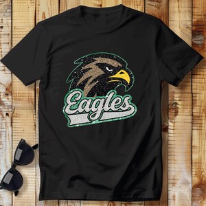 K&ouml;nnte beinhalten: Schwarzes T-Shirt mit einem glitzernden Adlerkopf-Design und dem Wort "Eagles" in einer passenden glitzernden Schriftart. Der Adler hat braune, schwarze und gelbe Akzente. Sonnenbrille liegt auf einer Holzoberfl&auml;che.