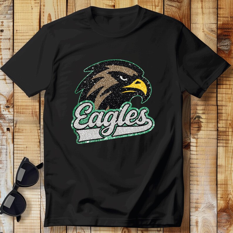 K&ouml;nnte beinhalten: Schwarzes T-Shirt mit einem glitzernden Adlerkopf-Design und dem Wort "Eagles" in einer passenden glitzernden Schriftart. Der Adler hat braune, schwarze und gelbe Akzente. Sonnenbrille liegt auf einer Holzoberfl&auml;che.