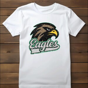 K&ouml;nnte beinhalten: Wei&szlig;es T-Shirt mit einem glitzernden Adlerkopf-Design mit braunen, schwarzen und gelben Akzenten. Das Wort "Eagles" wird in einer stilisierten Schriftart mit einem gr&uuml;nen und silbernen Rand angezeigt. Das Shirt besteht aus weichem, wei&szlig;em Stoff.