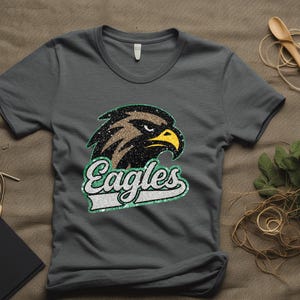 K&ouml;nnte beinhalten: Graues T-Shirt mit einem detaillierten Adlerkopf-Motiv mit braunen, schwarzen und gelben Akzenten. Das Wort "Eagles" wird in einer stilisierten Schriftart mit gr&uuml;n-wei&szlig;em Rand angezeigt. Das Shirt besteht aus einem weichen Material.