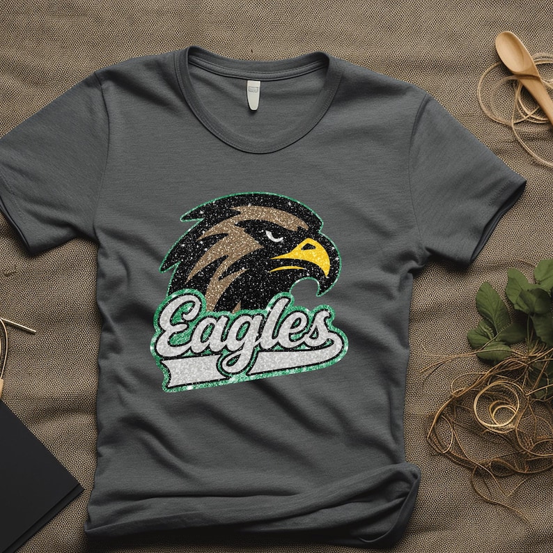 K&ouml;nnte beinhalten: Graues T-Shirt mit einem detaillierten Adlerkopf-Motiv mit braunen, schwarzen und gelben Akzenten. Das Wort "Eagles" wird in einer stilisierten Schriftart mit gr&uuml;n-wei&szlig;em Rand angezeigt. Das Shirt besteht aus einem weichen Material.