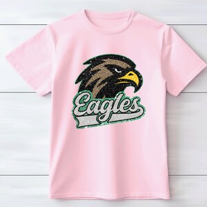 K&ouml;nnte beinhalten: Hellrosa T-Shirt mit einem glitzernden Adlerkopf-Design und dem Wort "Eagles" in einer passenden glitzernden Schriftart. Der Adler hat braune, schwarze und gelbe Akzente. Das T-Shirt besteht aus einem weichen Material.