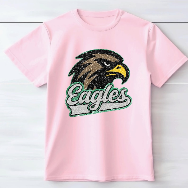 K&ouml;nnte beinhalten: Hellrosa T-Shirt mit einem glitzernden Adlerkopf-Design und dem Wort "Eagles" in einer passenden glitzernden Schriftart. Der Adler hat braune, schwarze und gelbe Akzente. Das T-Shirt besteht aus einem weichen Material.