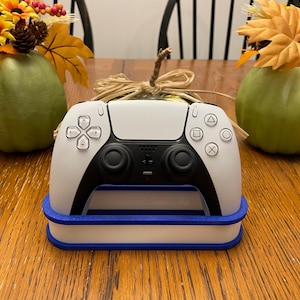 May include: A white and black video game controller sits on a white and blue stand. The controller is surrounded by fall-themed decorations, including pumpkins and autumn leaves. The controller has a variety of buttons and joysticks.