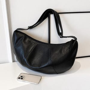 May include: A black crescent-shaped shoulder bag made of a smooth, slightly textured material with a single adjustable strap and a top zipper closure. A rectangular smartphone with a triple-lens camera system is positioned in the foreground.