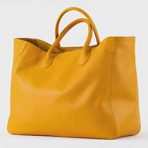 May include: A mustard yellow tote bag with a textured surface, featuring two rounded top handles attached to the main body. The bag has an open top design with a spacious interior compartment and a structured rectangular base, presented in a solid color.