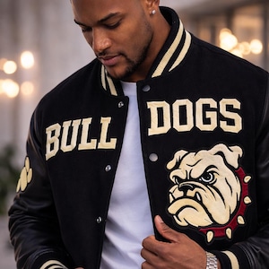 May include: Black varsity jacket with the words "BULL DOGS" in gold lettering. The jacket features a bulldog graphic and black leather sleeves. The collar and cuffs have gold and white stripes. The person is wearing a white t-shirt.