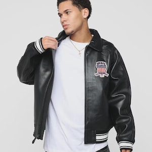 May include: Black leather jacket with a white t-shirt. The jacket features a patch with a red, white, and blue design and the text "AVIREX". The jacket has white and black striped cuffs and hem.