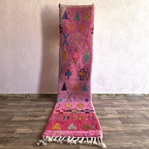 May include: A long, rectangular runner rug featuring a vibrant pink base with a high-pile texture. The surface is decorated with various small, colorful geometric shapes, including triangles, diamonds, and crosses in blue, yellow, and orange.